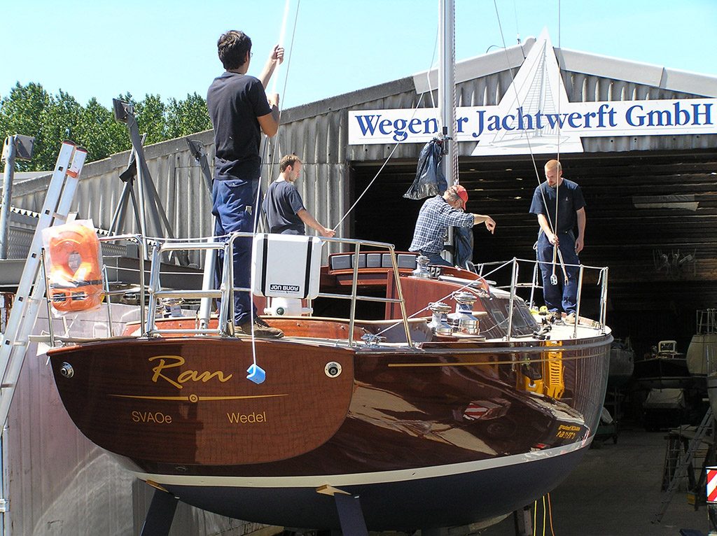 Vier Mitarbeiter riggen eine Mahagoni-Yacht vor der Halle mit dem Schild Wegener Jachtwerft GmbH